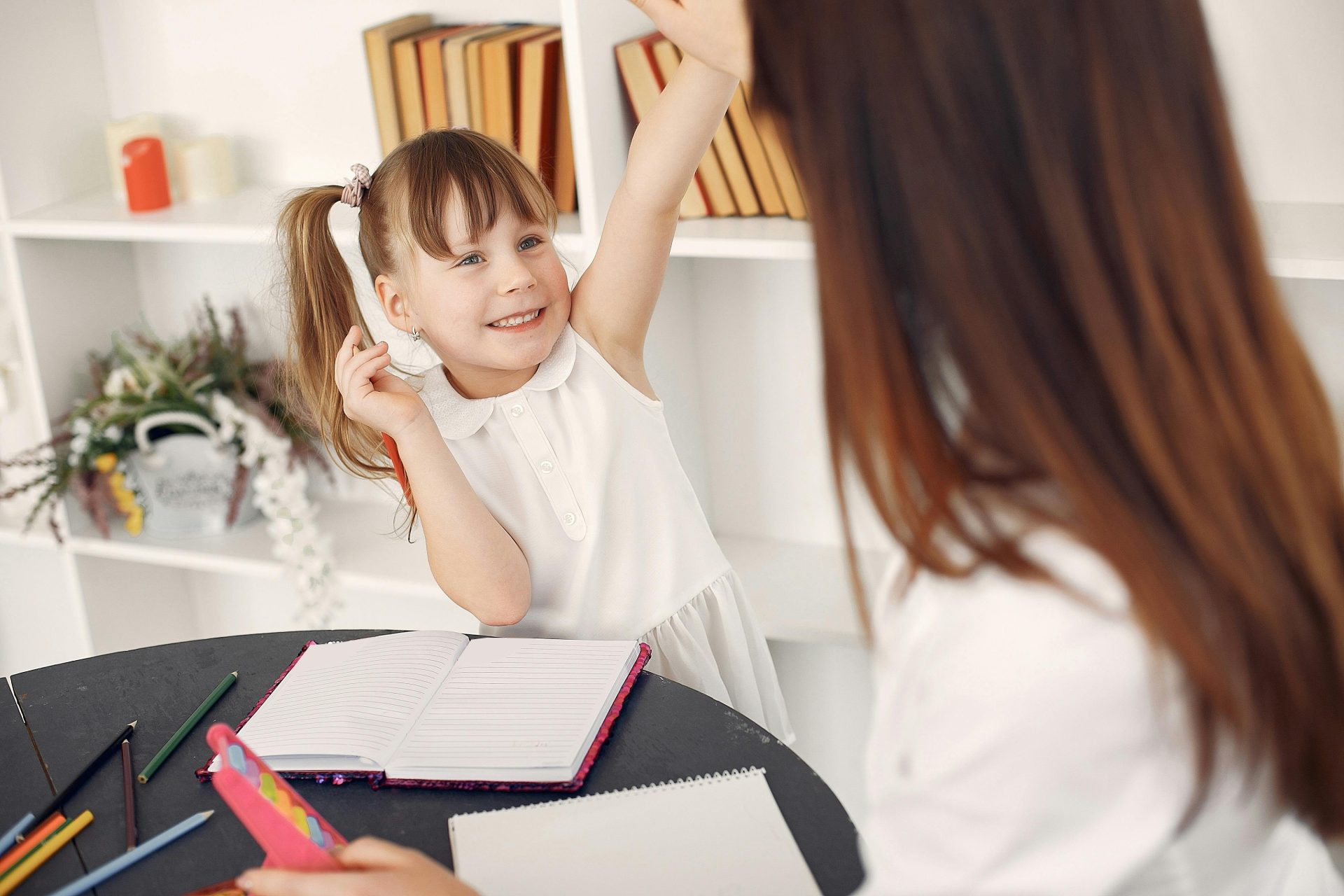 Smiling little girl in white dress and young female home teacher cheering each other with raised hands while sitting at table with notebook and pencils during home education against shelf with textbooks